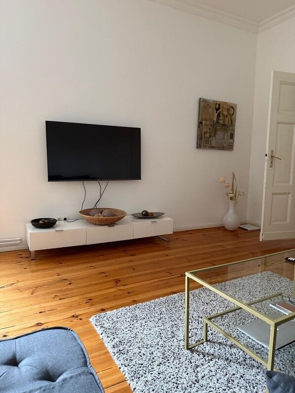 A modern living room with a flat-screen TV mounted on the wall, a white entertainment unit with decorative items, a glass coffee table, and a grey sofa.