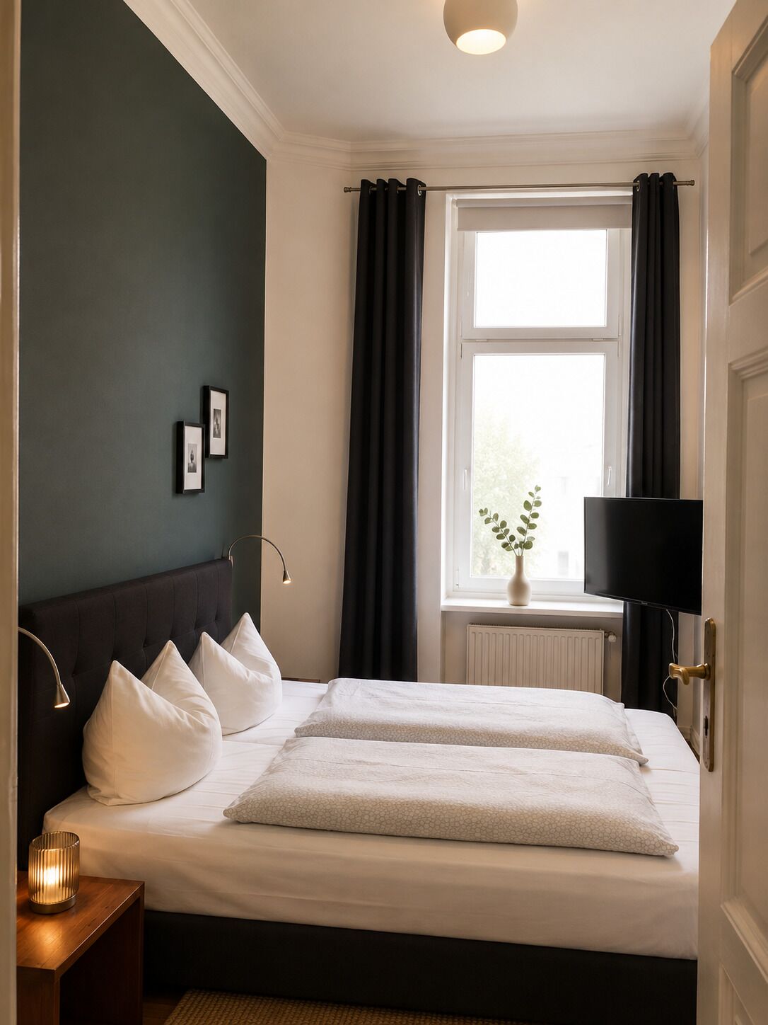 Une chambre à coucher avec un lit, des lampes de chevet, une fenêtre avec des rideaux, un radiateur et un téléviseur.