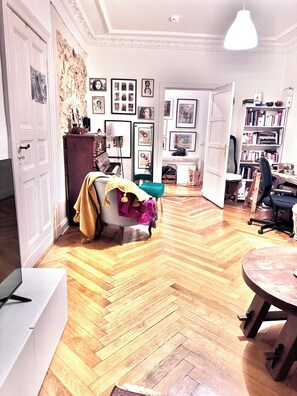 Een kamer met een visgraatparketvloer, een piano, een boekenkast en een bureau met een stoel.