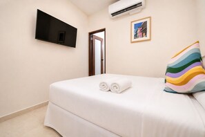 A hotel room with a bed, a flat-screen TV mounted on the wall, an air conditioning unit, and a door leading to another room.
