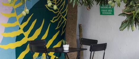 Una mesa y sillas bajo un árbol de hojas verdes y frutos inmaduros, junto a un muro con un letrero verde y un mural colorido.