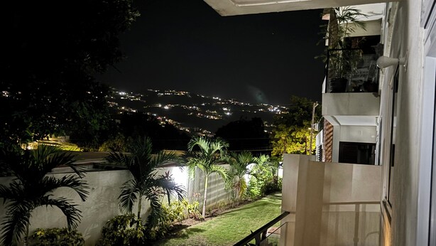 ’s Nachtzicht van een balkon met glazen hekwerk, met uitzicht op een verlichte tuin en een verafgelegen stadsbeeld.
