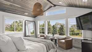A bedroom with a large bed, two chairs, a TV, and multiple windows offering a view of trees and a road.