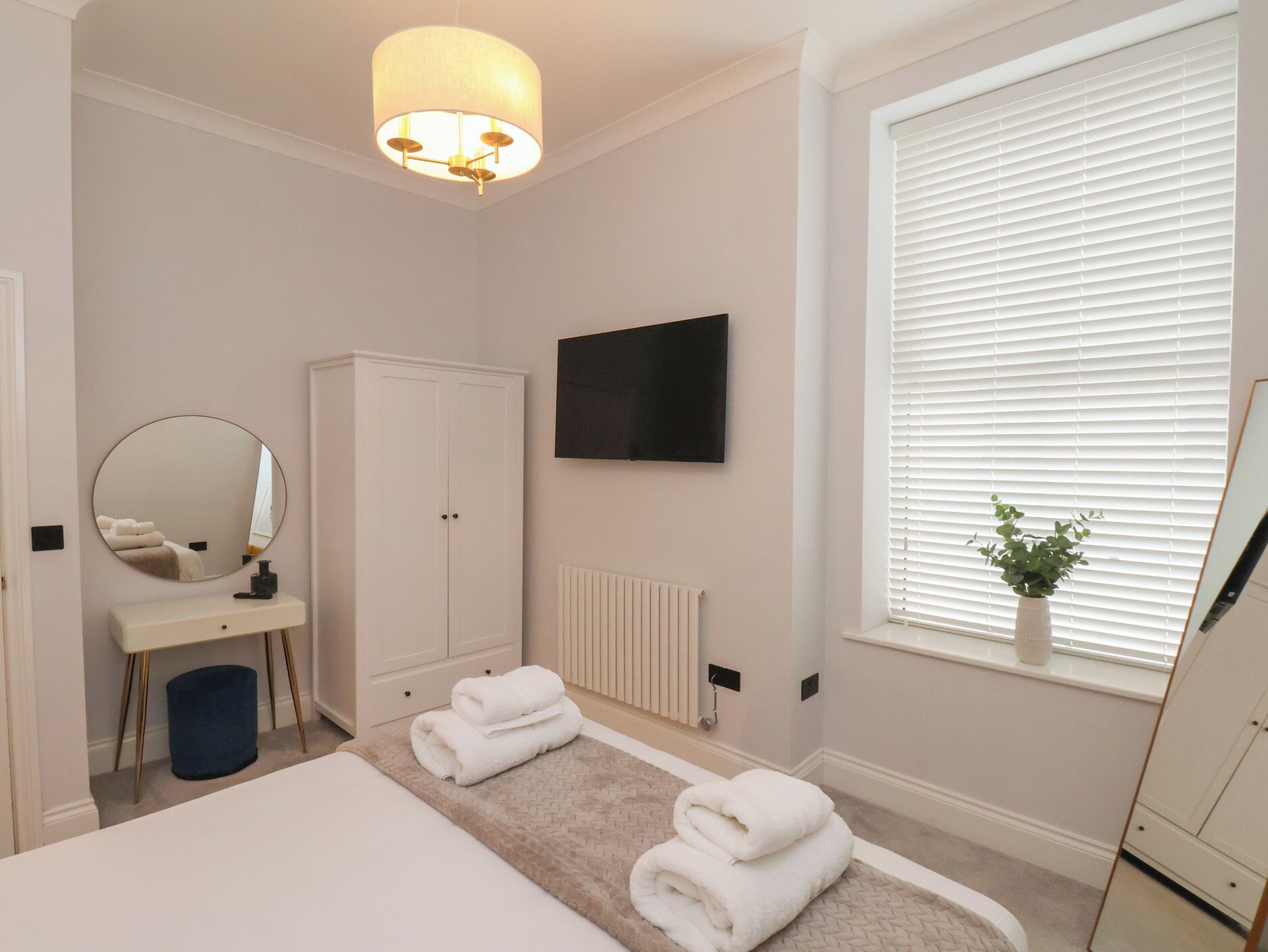 Een slaapkamer met een bed, een kledingkast, een spiegel, een radiator, een televisie en een raam met jaloezieën.