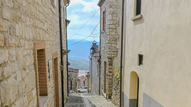 Une ruelle étroite pavée, bordée de bâtiments anciens en pierre, dotés d’arcades et de petites fenêtres.