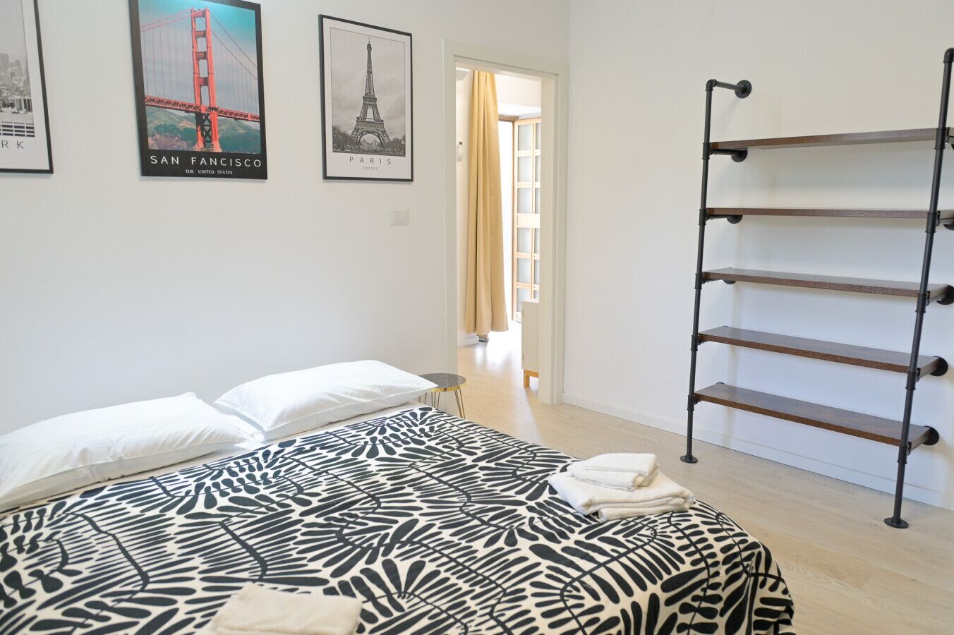 Ein Schlafzimmer mit einem Bett, einer schwarz-weiß gemusterten Tagesdecke, einem schwarzen Metalltrockner und zwei gerahmten Bildern der Golden Gate Bridge und des Eiffelturms.