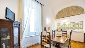 Un comedor con una mesa de madera, sillas, un televisor sobre un mueble bajo y una ventana con cortinas.