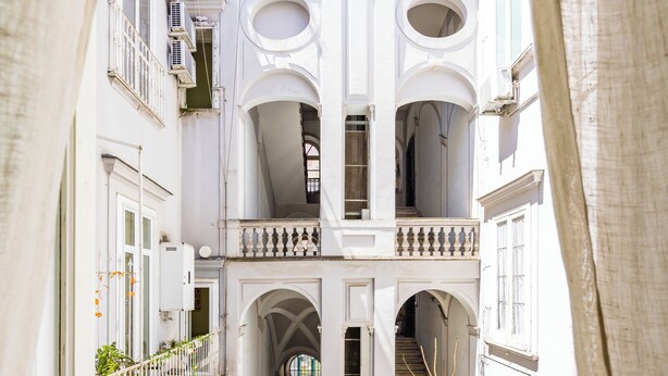 Un patio estrecho con una escalera que lleva a un balcón en el segundo piso, rodeado de edificios blancos con balcones y arcos.