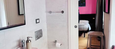 Un baño con inodoro, lavamanos y espejo. Hay una zona de ducha con un perchero y un dispensador de papel higiénico. El baño tiene una decoración en tonos rosa y amarillo.