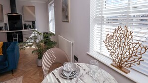 A modern living room with a blue sofa, a dining table set for two, and a kitchen in the background. - The Salisbury - 1 Bedroom Deluxe Apartment (Scarborough)