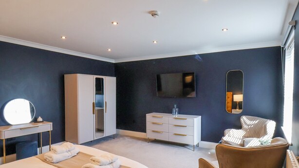 A modern hotel room with a bed, a vanity with a mirror, a dresser, a television, and a chair. - The Salisbury - 1 Bedroom Deluxe Apartment (Scarborough)