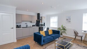 A modern living room with a blue sofa, a glass coffee table, and a kitchen area with white cabinets and a black range hood. - The Salisbury - 1 Bedroom Deluxe Apartment (Scarborough)
