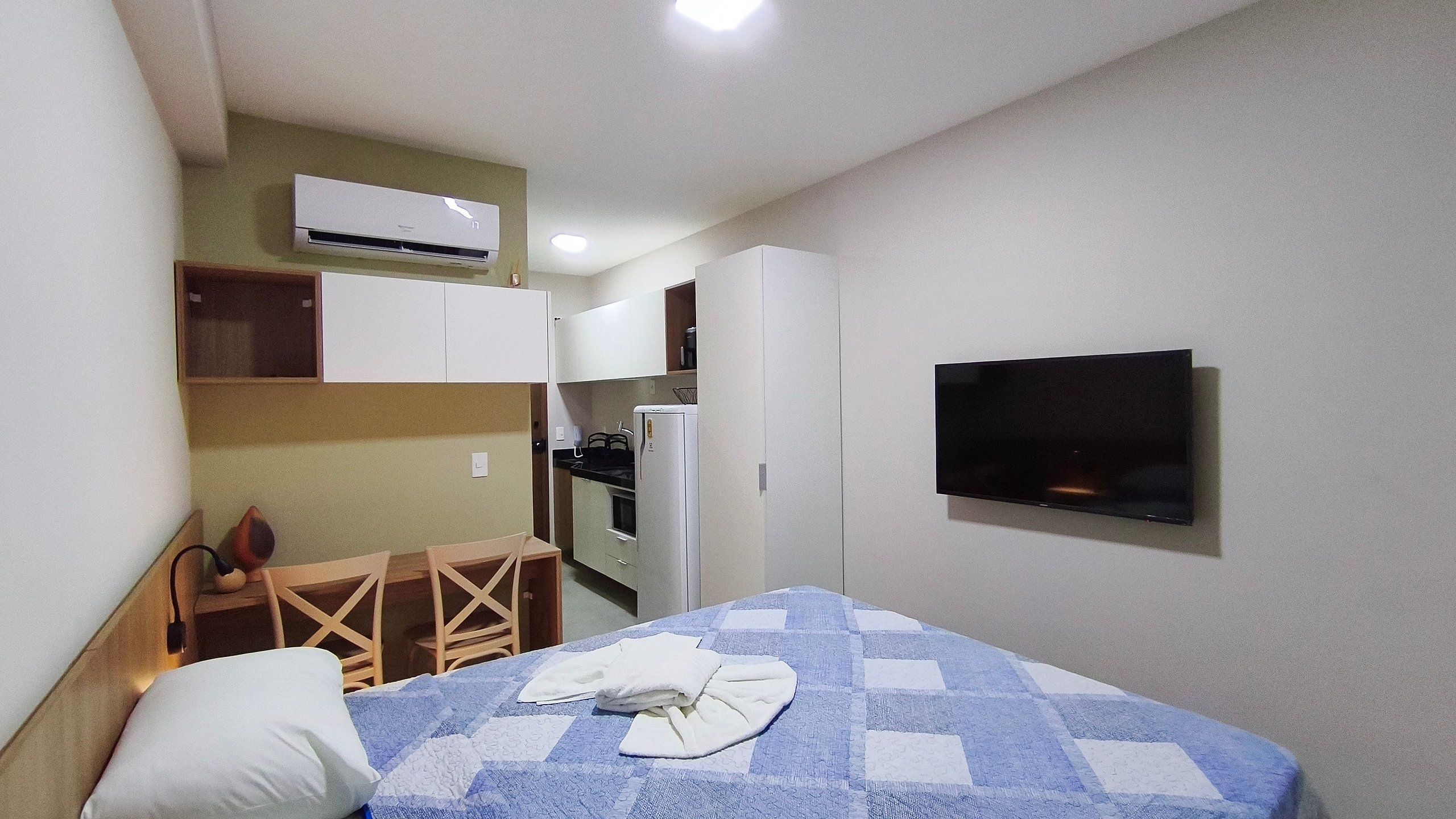 Quarto de hotel pequeno com uma cama, uma televisão na parede, uma cozinha compacta com geladeira e fogão, e uma cadeira de madeira.
