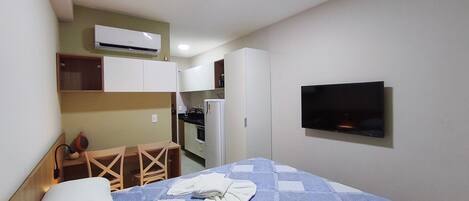 Una habitación de hotel pequeña con una cama, un televisor fijado a la pared, una kitchenette con refrigerador y estufa, y una silla de madera.