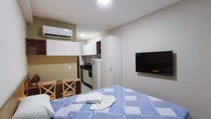 A small hotel room with a bed, a TV mounted on the wall, a kitchenette with a fridge and stove, and a wooden chair.