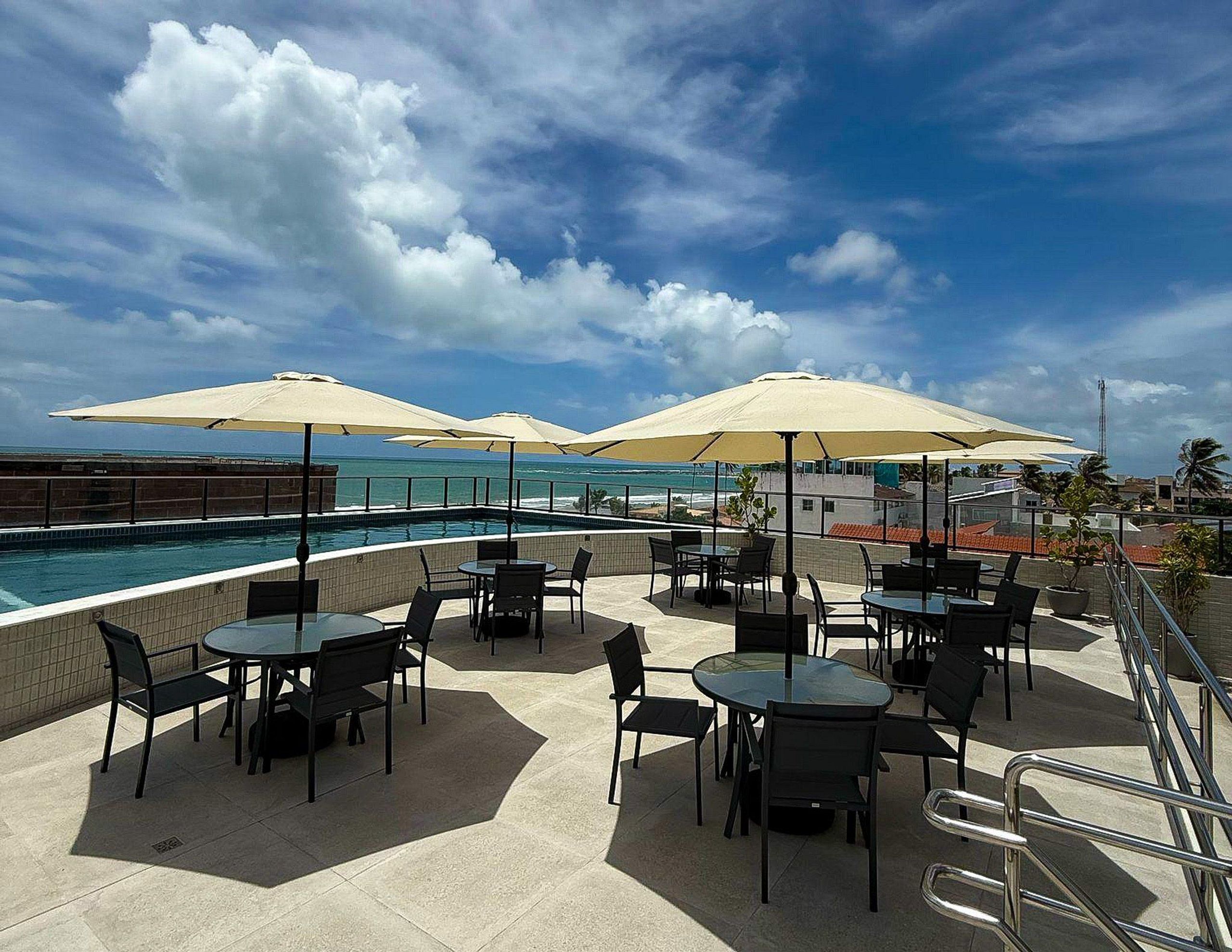 A rooftop pool area with tables and chairs, umbrellas, and a view of the ocean.