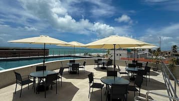 Uma área de piscina no terraço com mesas, cadeiras, guarda-sóis e vista para o oceano.