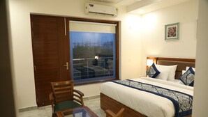 A hotel room with a bed, a table with chairs, a window with a view, and an air conditioning unit. - Hotel Sandal Tree Express (Noida)