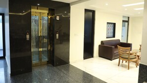 A modern lobby with a black elevator, a brown sofa, and a wooden table with chairs. - Hotel Sandal Tree Express (Noida)