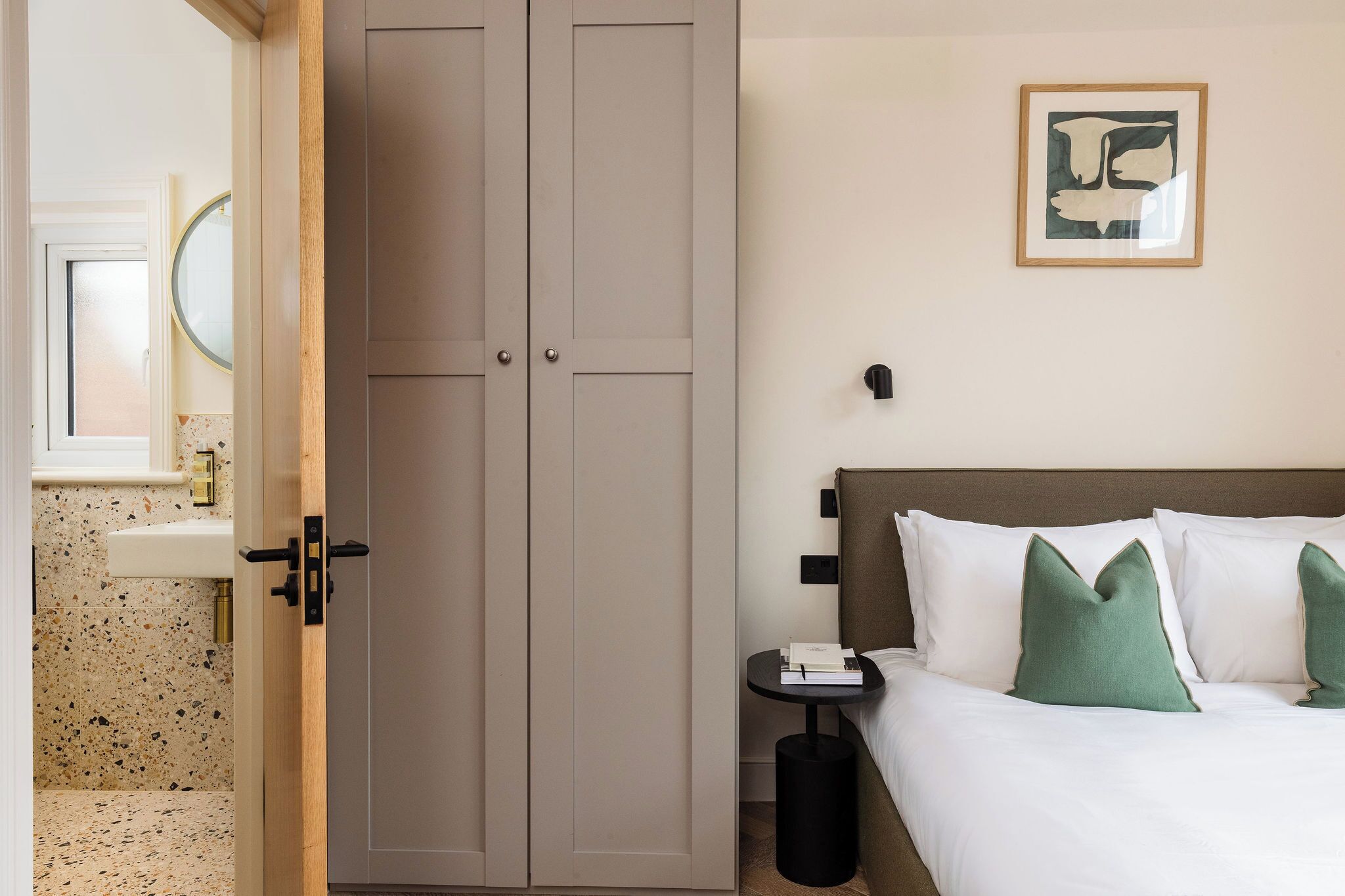 A hotel room with a bed, a nightstand, a wardrobe, a bathroom with a sink, and a framed picture on the wall.