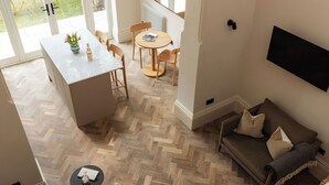 A modern living space with a herringbone floor pattern, a central island with chairs, a sofa with cushions, and a flat-screen TV mounted on the wall. - FiveM West Hampstead - South (London)