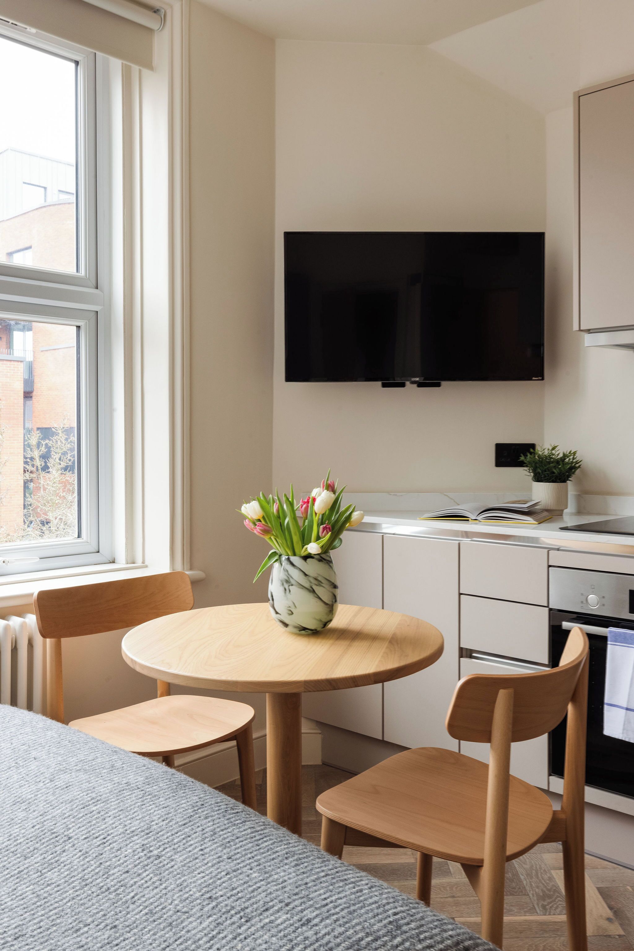 A modern kitchen with a round dining table, two chairs, a flat-screen TV mounted above the counter, and a potted plant on the counter.