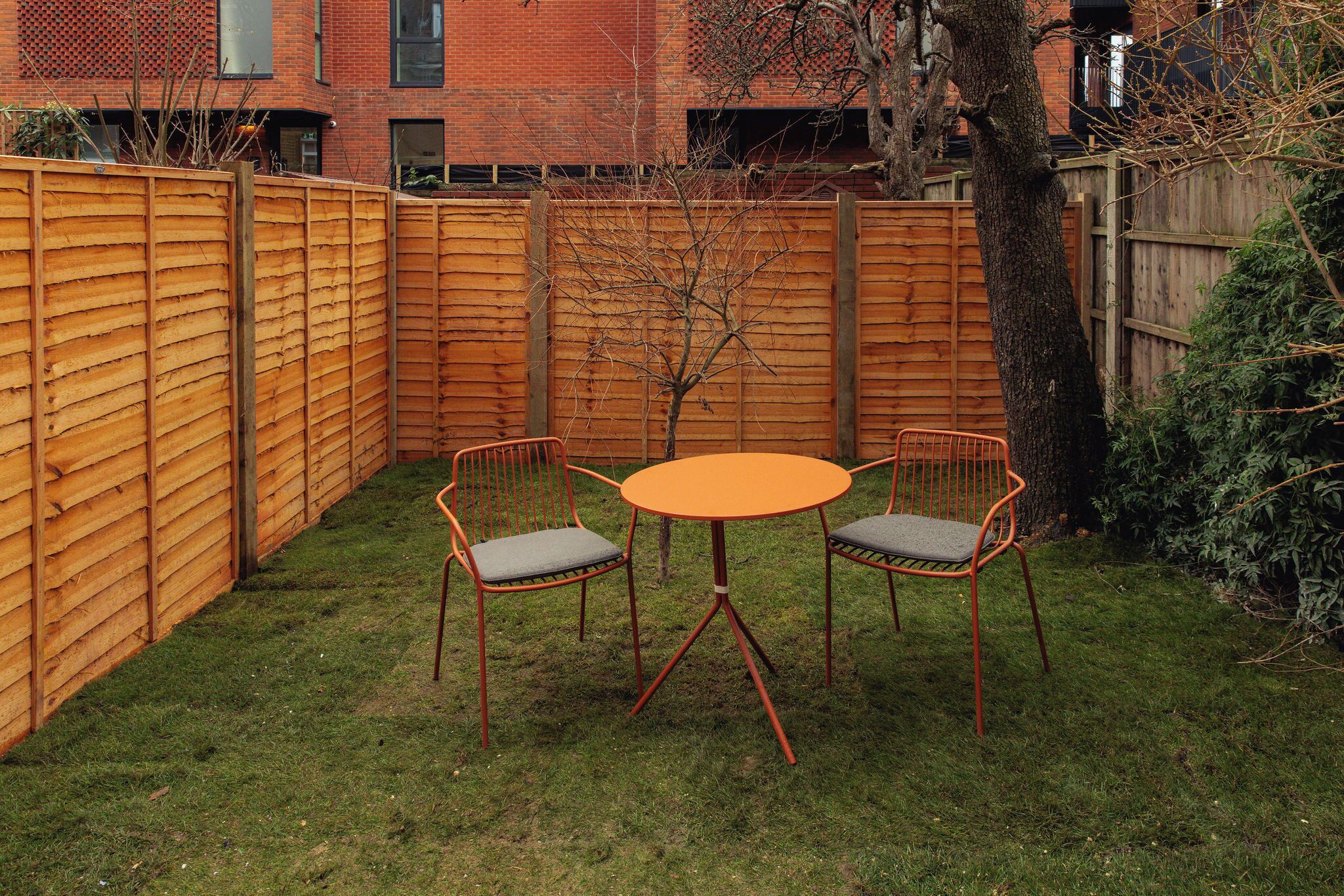 Un pequeño espacio al aire libre con una mesa redonda y cuatro sillas, rodeado por una valla de madera y un árbol.