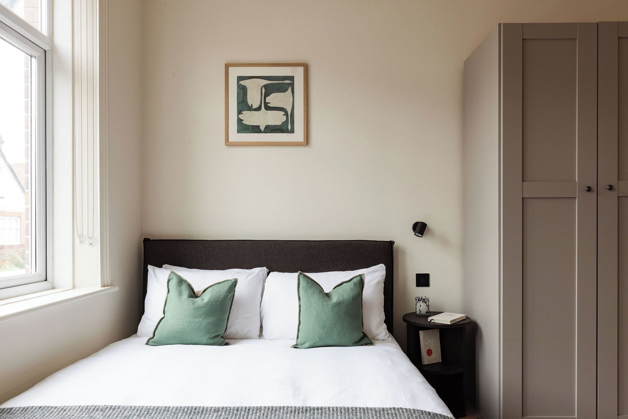A neatly made bed with white and green pillows, a framed artwork on the wall, a bedside table with a book, and a wardrobe.