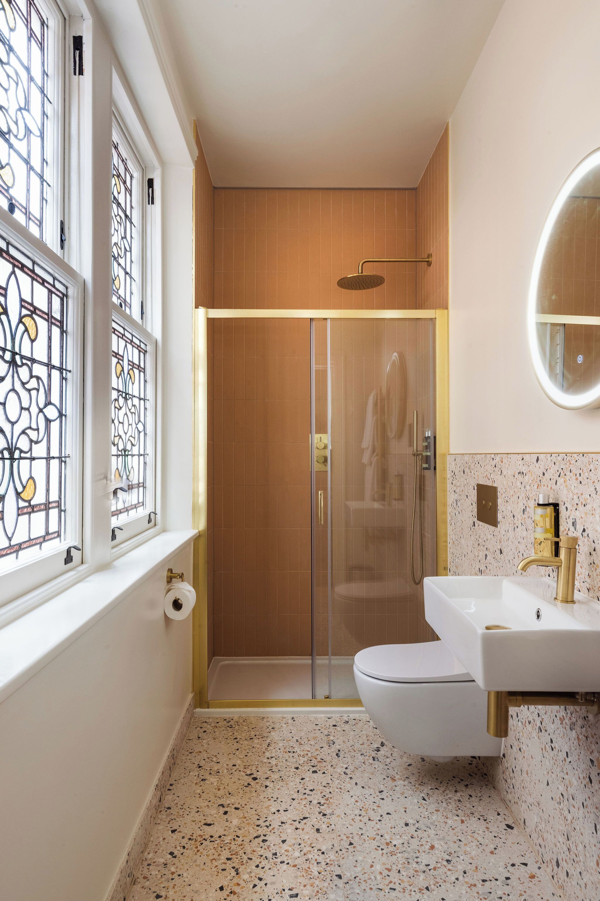 A modern bathroom with a glass-enclosed shower, a white toilet, a sink with a gold faucet, and a window with stained glass.