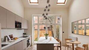 A modern kitchen with a central island, a dining area with wooden chairs, and large windows offering a view of the outside. - FiveM West Hampstead - South (London)