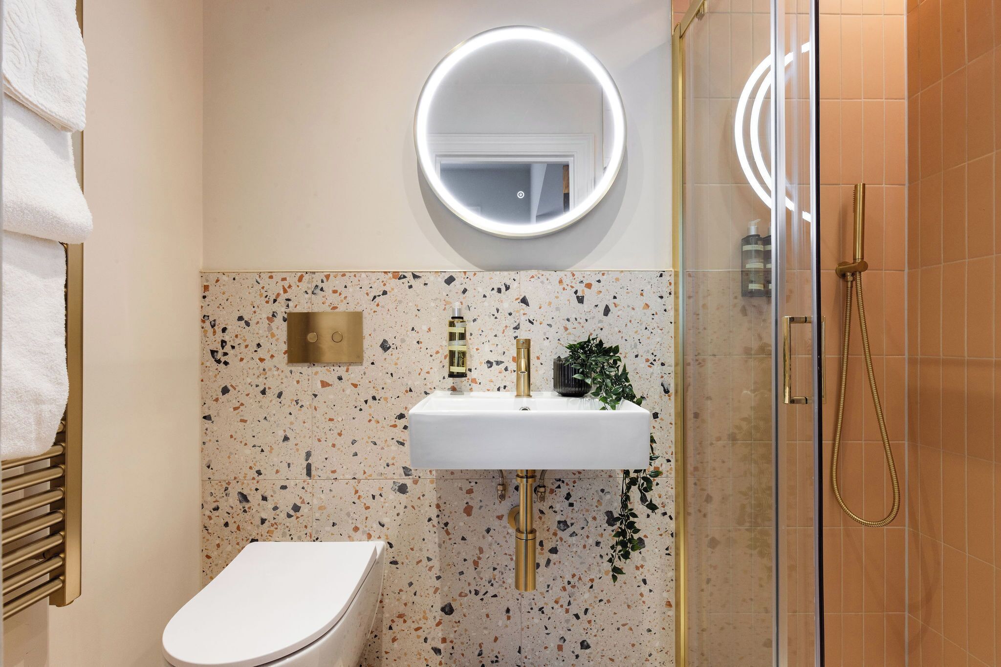 A bathroom with a white toilet, a sink with a gold faucet, a round mirror with a light, and a shower with a gold hose.