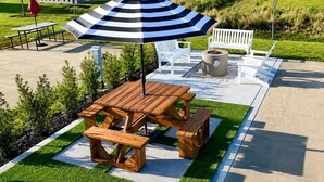 Un patio al aire libre con una mesa de picnic de madera, bancos, un paraguas a rayas y muebles de exterior blancos.