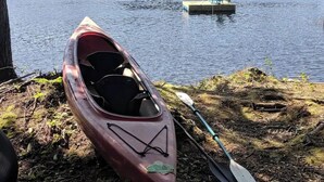 Un canoë est amarré à un quai, une pagaie est posée à proximité, un petit bateau se trouve à l’arrière-plan et une côte boisée borde l’eau.