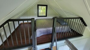 Une petite chambre mansardée avec un plancher en bois, un lit doté d’un matelas gris, une fenêtre donnant sur la verdure et une rambarde noire.