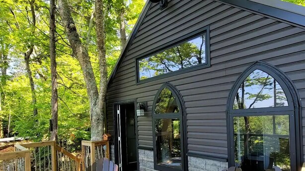 Une terrasse en bois avec deux fauteuils Adirondack, une cheminée en pierre et une maison aux bardages sombres et aux grandes fenêtres.