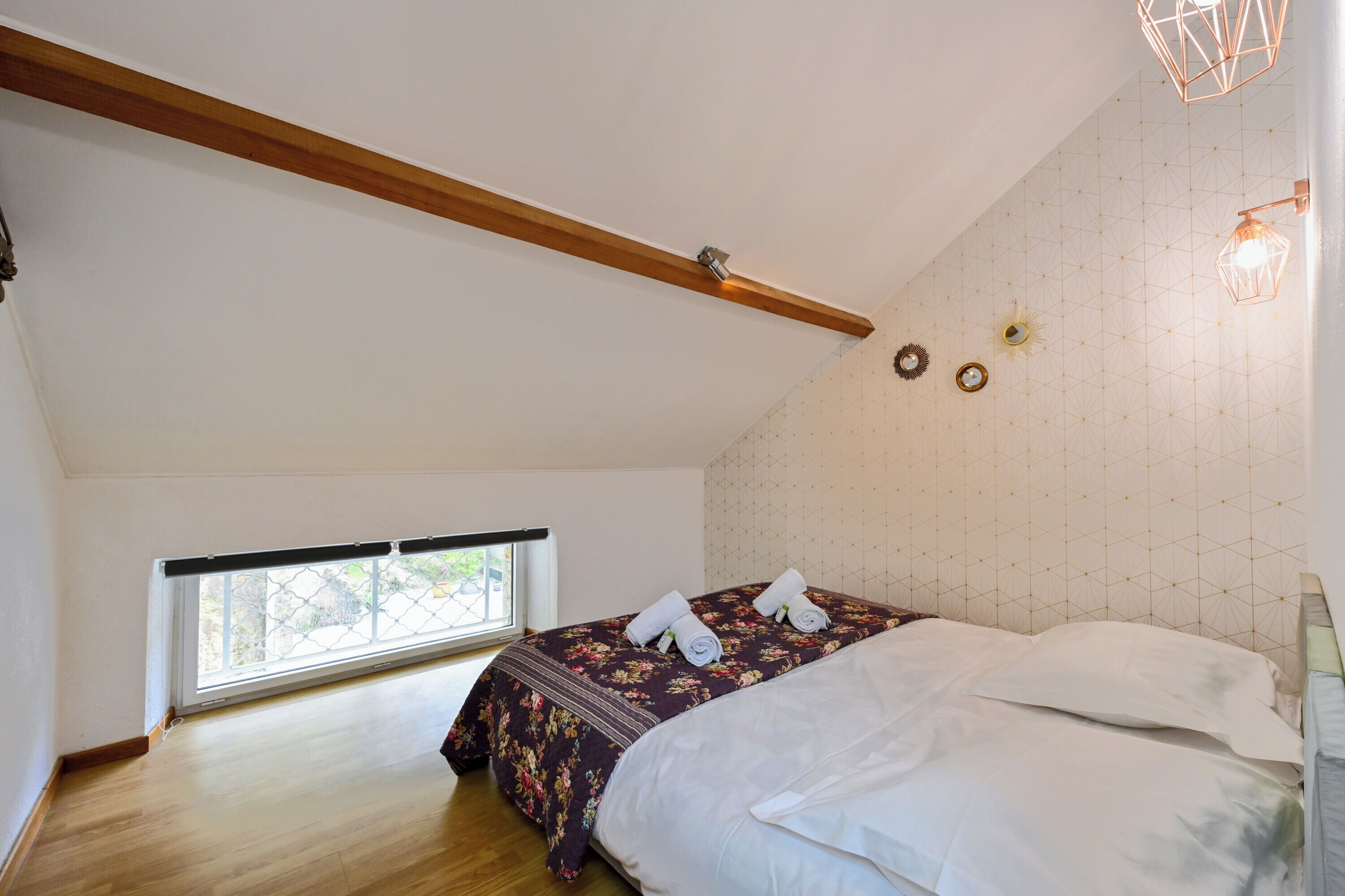 A bedroom with a wooden ceiling beam, a bed with white linens, a floral-patterned bedspread, and a geometric-patterned wall.