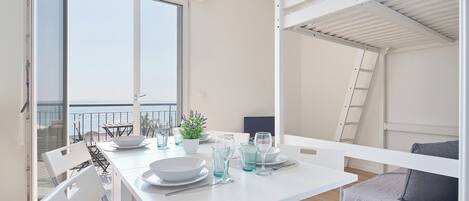 A modern, well-lit dining area with a white table set for four, a balcony with a view, and a bunk bed with a ladder.
