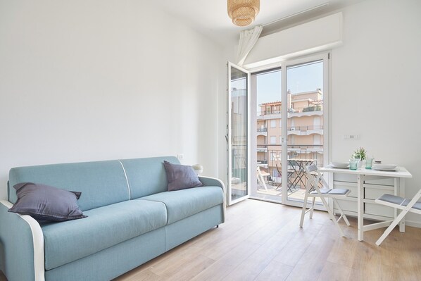 Ein modernes Wohnzimmer mit einem hellblauen Sofa, einem kleinen weißen Tisch und einem Balkon mit Blick auf Gebäude.