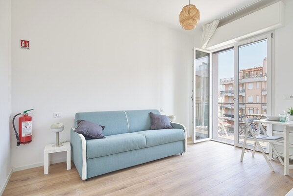 A modern living room with a light blue sofa, a small white side table, a floor lamp, and a glass door leading to an outdoor balcony.