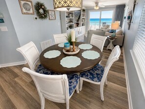 A dining area with a round table, white chairs, and a view of the ocean through large windows. - Beachfront Bliss! Gulf Waves at The Palms #201 (Okaloosa Island)