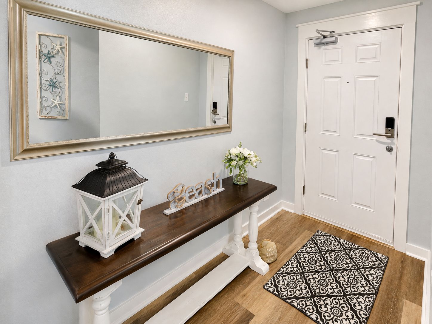 A hallway with a dark wooden console table, a white door, a large mirror, and a patterned rug.