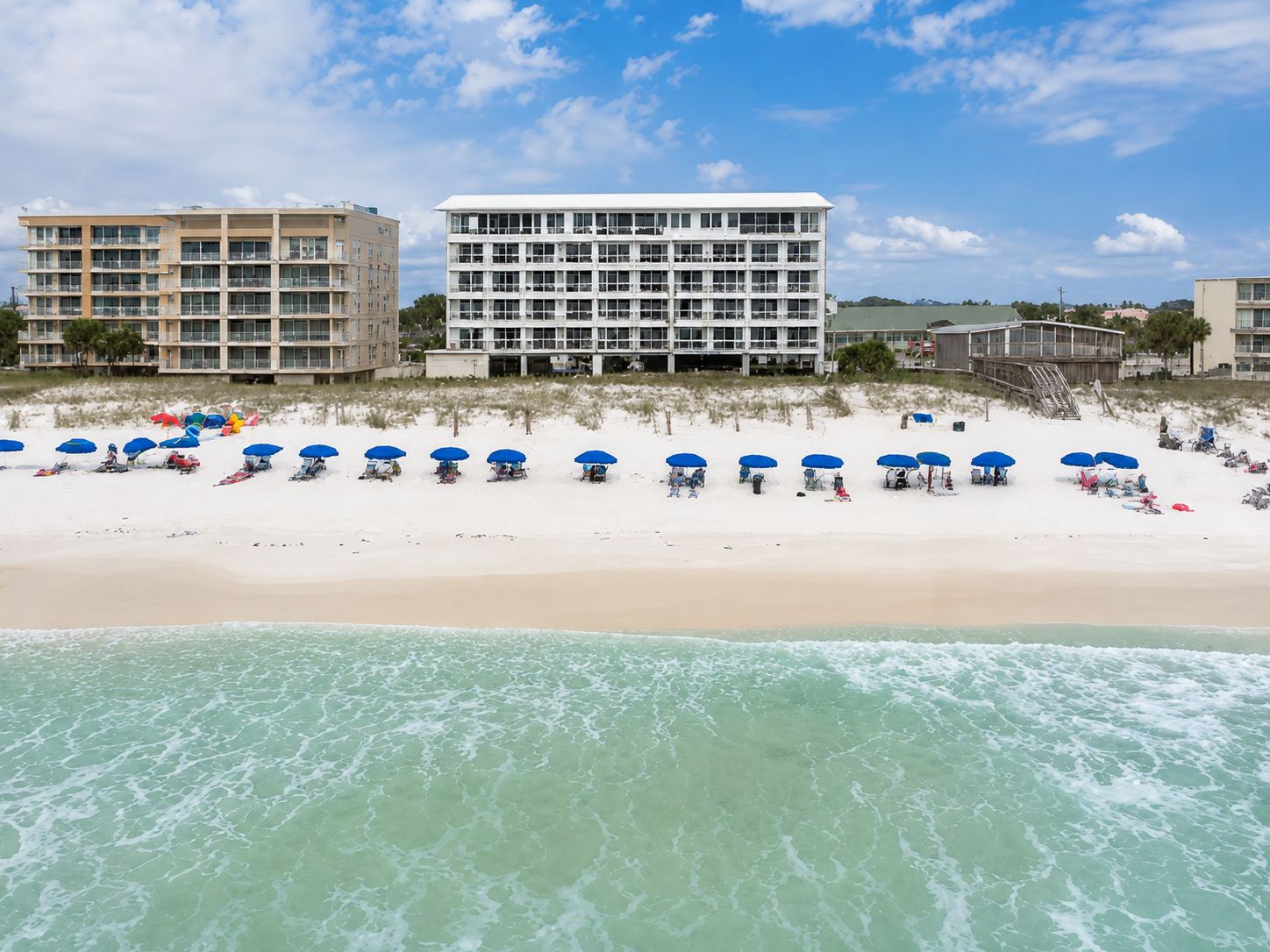 A beach with rows of blue umbrellas, a building with multiple floors, and a clear sky.
