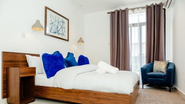 A modern bedroom with a wooden bed, blue and white pillows, a blue chair, and a large window with curtains. - PREMIUM SUITE (Accra)