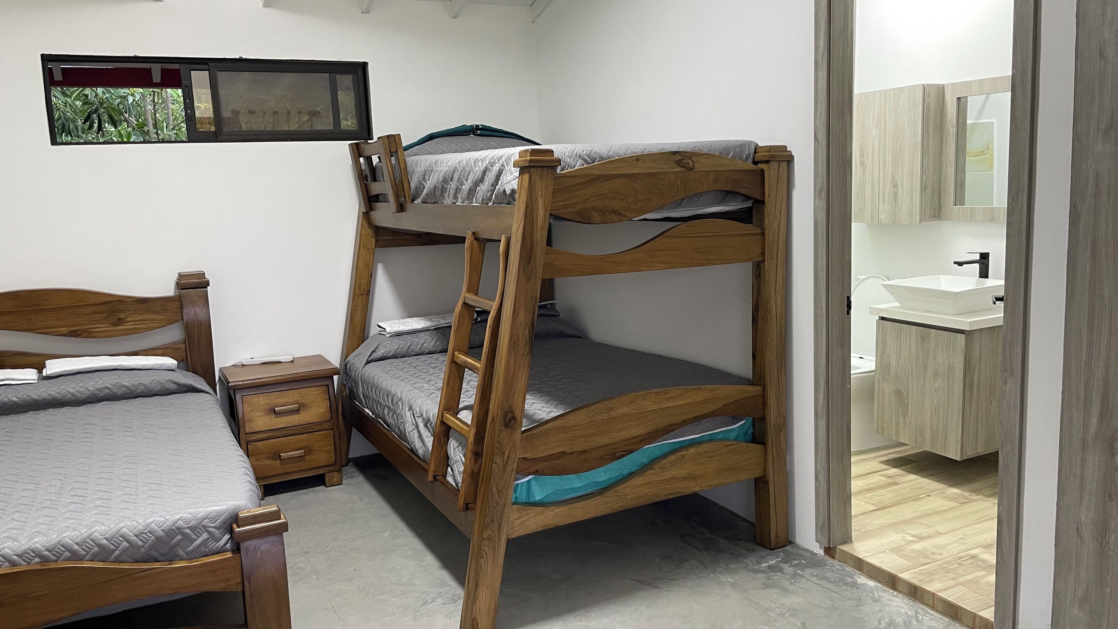 A bunk bed with a wooden frame, a bedside table with drawers, a small bedside lamp, and a window with a view of trees.