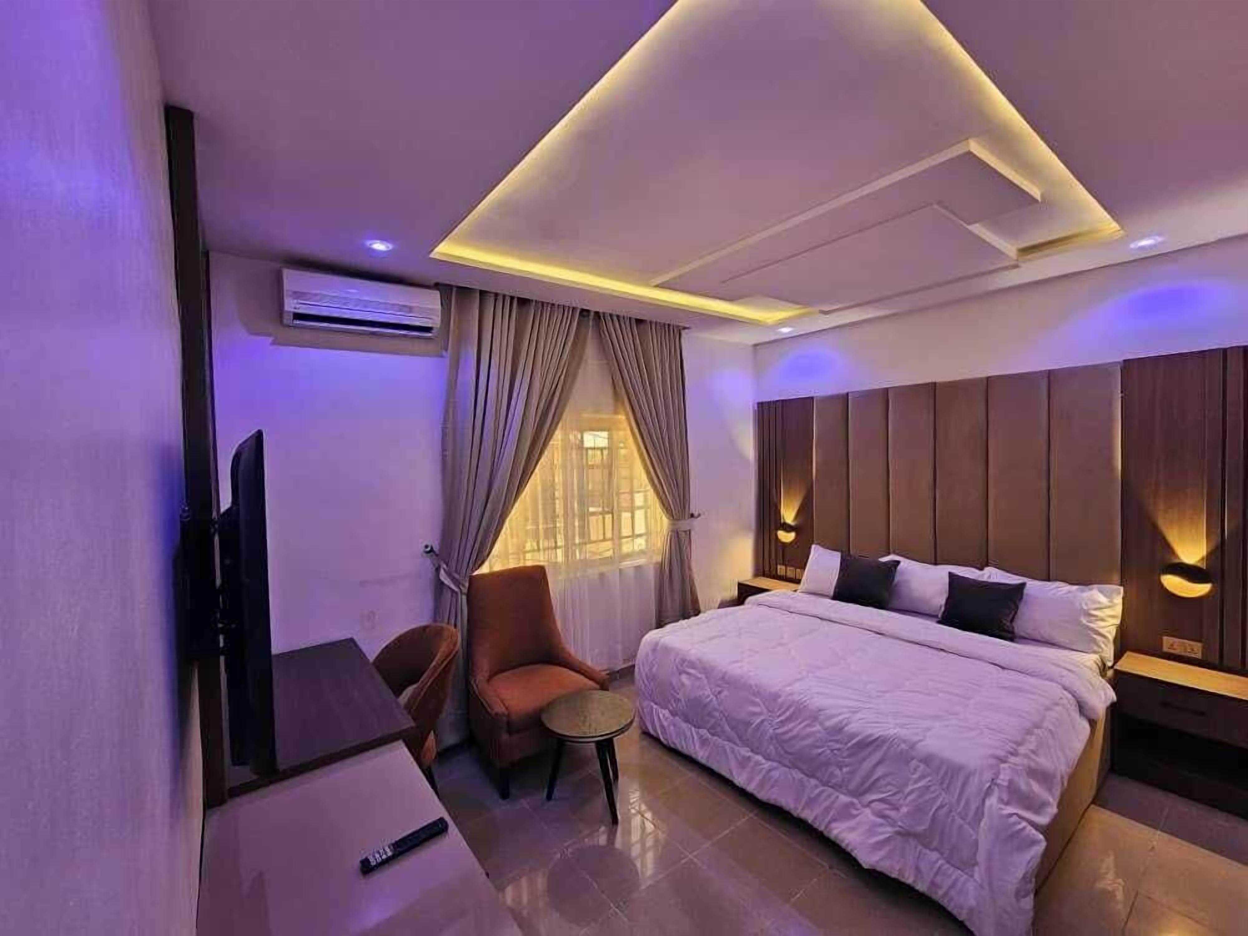 A modern hotel room with a large bed, a desk, and a chair. There is a TV on the wall and a window with curtains.