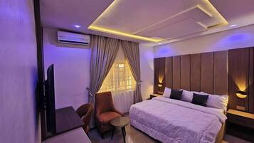 A modern hotel room with a large bed, a desk, and a chair. There is a TV on the wall and a window with curtains.