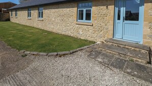 A stone building with a blue door and windows, a gravel driveway, and a grassy lawn. - Bower Cottage (Beaminster)