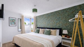A hotel room with a large bed, a bedside table with a lamp, a wooden ladder, and a window with curtains.