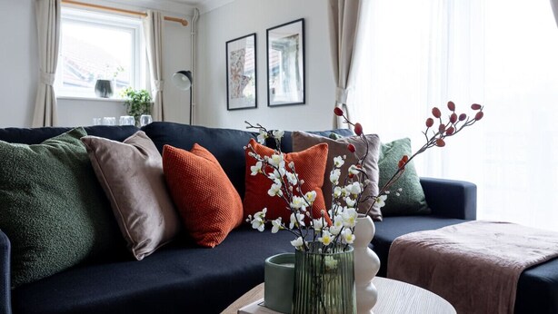A living room with a dark blue sofa, multiple cushions, a vase with flowers, and a window with curtains.