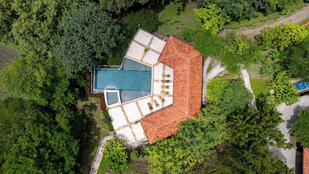 Eine Vogelperspektive zeigt ein Anwesen mit Schwimmbad, einer Terrasse mit Sitzgelegenheiten und einem Gebäude mit rotem Dach, umgeben von üppiger Vegetation.